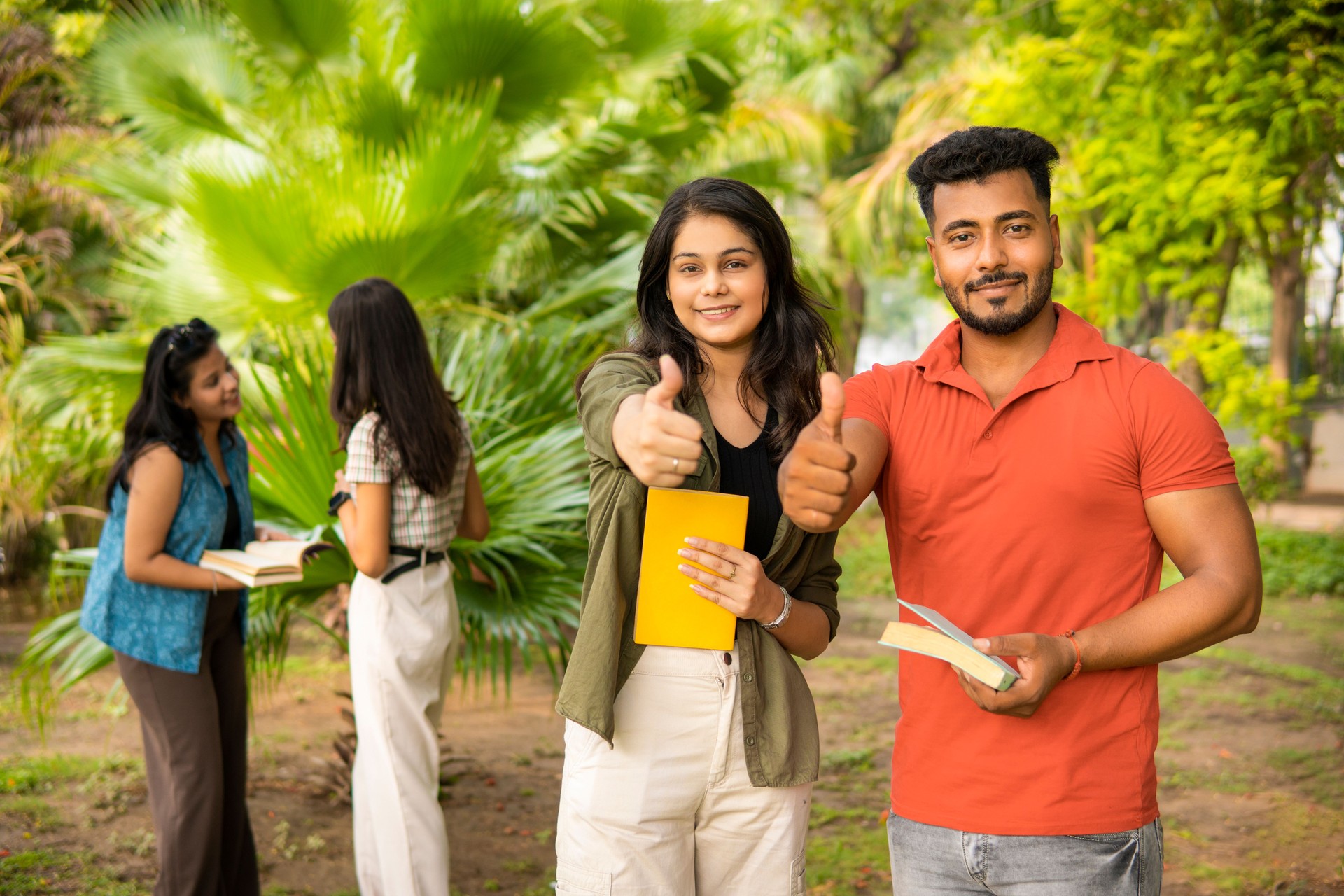 Portrait of two young students holding a book and showing thumbs up to the camera on university campus.